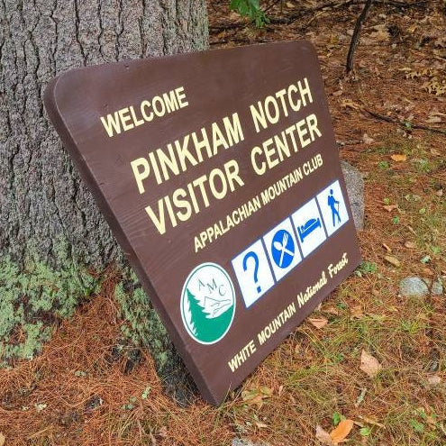 A brown wooden sign reading 'WELCOME PINKHAM NOTCH VISITOR CENTER' with white text and small blue and green icons, leaning against a large tree in a wooded outdoor setting.