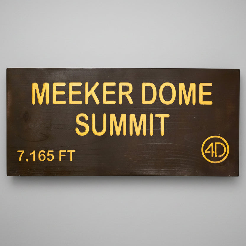 A rectangular dark brown wooden sign with yellow lettering that reads 'MEEKER DOME SUMMIT' and '7,165 FT', plus a circular '4D' logo, mounted against a dark wood plank wall.