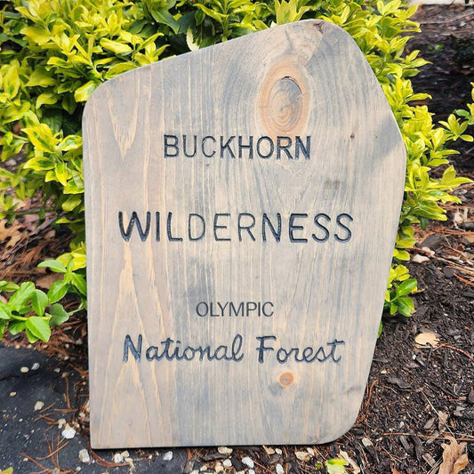 A rustic, handcrafted wooden sign with a weathered gray-brown finish, featuring the text 'BUCKHORN WILDERNESS OLYMPIC National Forest' in black lettering. The sign is photographed outdoors in a garden bed surrounded by green shrubs and mulch.