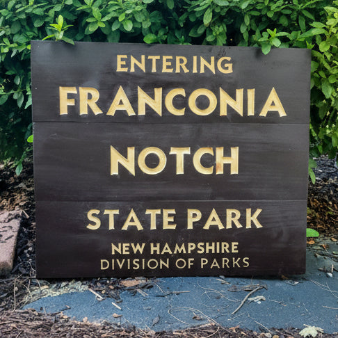 A dark brown wooden sign with raised gold lettering that reads 'ENTERING FRANCONIA NOTCH STATE PARK NEW HAMPSHIRE DIVISION OF PARKS,' photographed outdoors in front of green foliage.