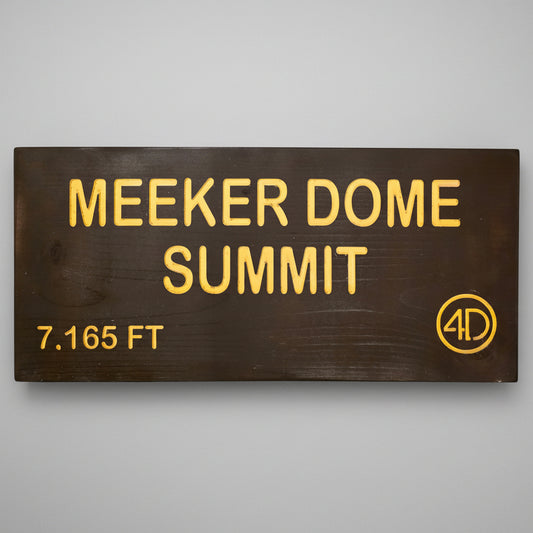 A rectangular dark brown wooden sign with yellow lettering that reads 'MEEKER DOME SUMMIT' and '7,165 FT', plus a circular '4D' logo, mounted against a dark wood plank wall.