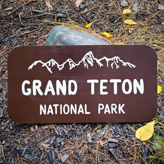 A dark brown wooden sign for Grand Teton National Park with white text and mountain graphics, placed outdoors on mulch with a rock behind it.